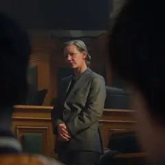 Sandra Hüller, con traje gris, se mantiene firme en una sala de audiencias, con las manos entrelazadas, mientras la cámara se enfoca en su rostro pensativo.