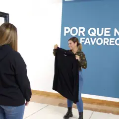 Una mujer sonríe mientras sostiene una prenda en tonos de negro frente a otra mujer que mira en un espejo.