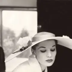 Mujer con sombrero de ala ancha y rosa, estilo Helmut Newton. Un hombre en traje con pajarita al fondo.