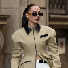 Mujer con gafas de sol oscuras y pendientes dorados, vistiendo una chaqueta beige con cuello negro y cinturón. Un look muy chic.