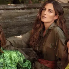 Belen Corsini y dos mujeres más, vestidas con ropa de moda, posan sentadas en un banco de madera rústico.