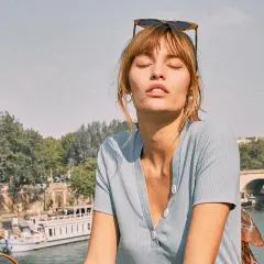 Mujer con gafas de sol en la cabeza y un pañuelo con estampado vichy, disfrutando del sol junto a un río con un barco y un puente al fondo.