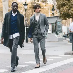Dos hombres cruzan una calle, uno con abrigo y jersey azul, el otro con traje gris. Representan la moda slow, atemporal y consciente.