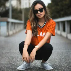 Chica con gafas de sol y camiseta naranja a rayas, agachada en un puente. Su atuendo es muy comfy y moderno.