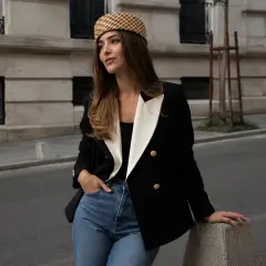 Mujer con un elegante bléiser negro y blanco, jeans y sombrero a cuadros, apoyada en un poste de concreto en la calle.