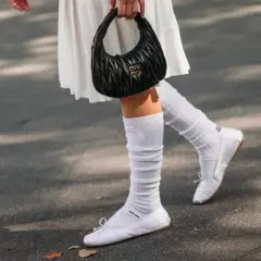 Piernas con bailarinas blancas y calcetines blancos arrugados, un bolso negro Miu Miu en la mano.