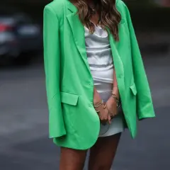 Chaqueta de **verde color** vibrante sobre un vestido blanco satinado. Brazaletes dorados adornan sus muñecas.