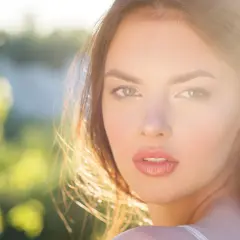 Una joven con un brillo natural, un verdadero glow up, mira a la cámara con el sol iluminando su rostro y el fondo verde desenfocado.