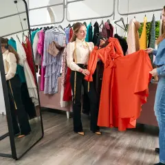 Dos mujeres discuten el outfit significado de un vestido naranja vibrante en una boutique.
