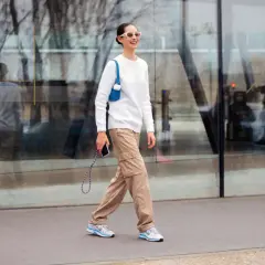 Mujer sonriente camina con un suéter blanco y un pantalón beige de carga, luciendo gafas de sol blancas y un bolso azul.