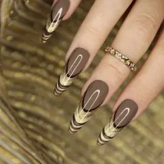 Uñas marrones tendencia con diseño de líneas doradas y brillo. Un anillo dorado adorna el dedo.