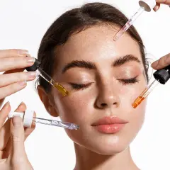 Manos aplican sueros con goteros en el rostro de una mujer. El brillo en su piel sugiere el poder de la vitamina C para la cara.