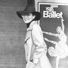 Elsa Peretti, con sombrero y gafas, sonríe frente a un cartel del New York City Ballet.