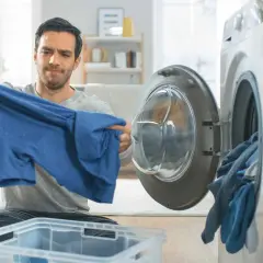 Hombre mira con preocupación una camiseta azul, parece que la viscosa encoge. La lavadora está abierta con ropa dentro.