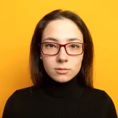 Joven con gafas de cara alargada y estrecha, pecas y jersey negro, sobre fondo amarillo.