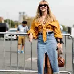 Mujer sonriente con un outfit falda vaquera larga, top amarillo anudado y bolso marrón.