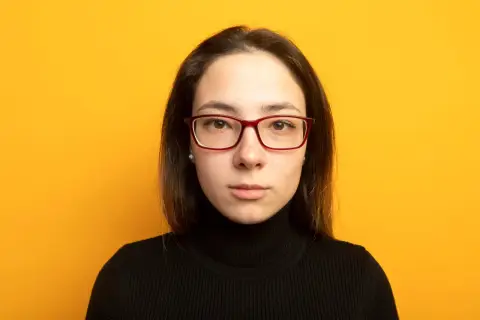 Joven con gafas de cara alargada y estrecha, pecas y jersey negro, sobre fondo amarillo.