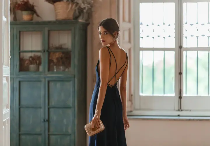 Mujer con un elegante vestido azul marino, perfecto para combinar en una boda. Lleva un bolso dorado y mira por encima del hombro.