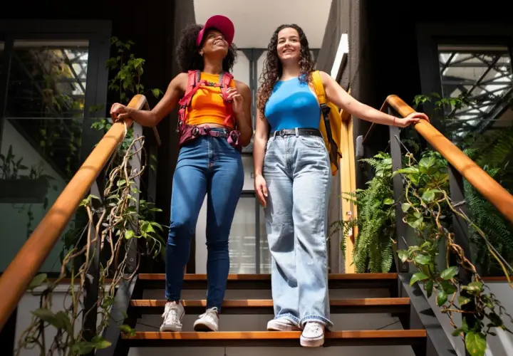 Dos amigas con estilo y2k, una con top naranja y gorra rosa, la otra con top azul, bajan unas escaleras rodeadas de plantas.