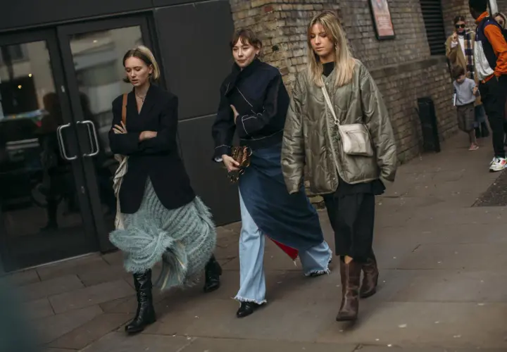 Tres mujeres caminan por la calle, cada una con un estilo único. La de la izquierda lleva unas **botas** negras altas y una falda de flecos. La del medio, un conjunto vaquero y unas botas negras. La de la derecha, unas botas marrones y una chaqueta aco...