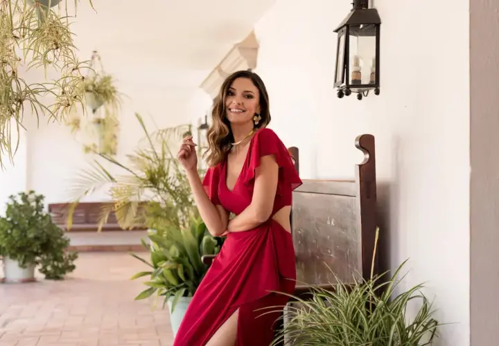 Mujer sonriente con un elegante outfit bautizo rojo, con detalles de corte y volantes, posando entre plantas y un banco de madera.