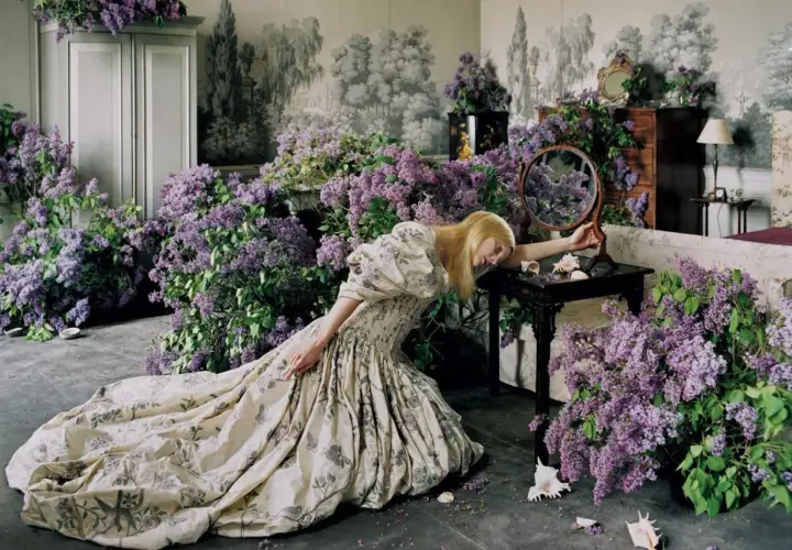 Una mujer rubia con un vestido estampado cae sobre un montón de lilas, evocando la estética onírica de Tim Walker.