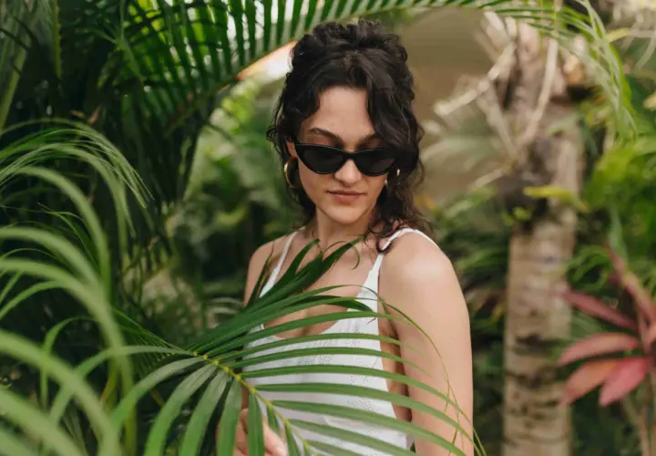 Joven con gafas de sol y traje de baño blanco entre hojas verdes, un ejemplo de moda ecológica.