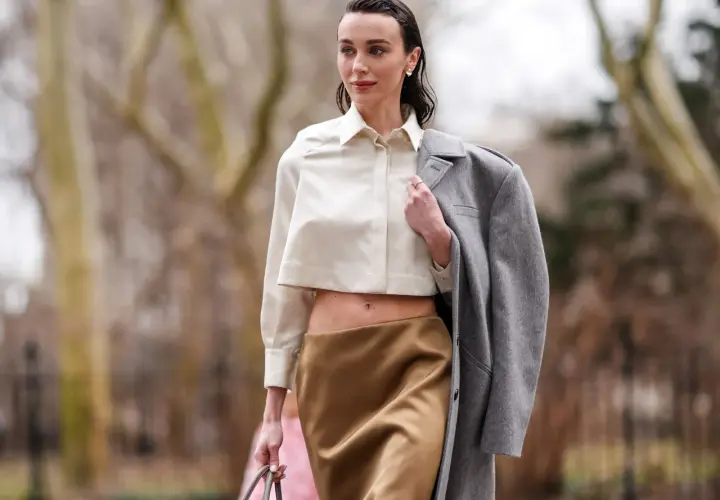 Mujer con blusa corta y falda satinada color caramelo, abrigo gris colgado del hombro.