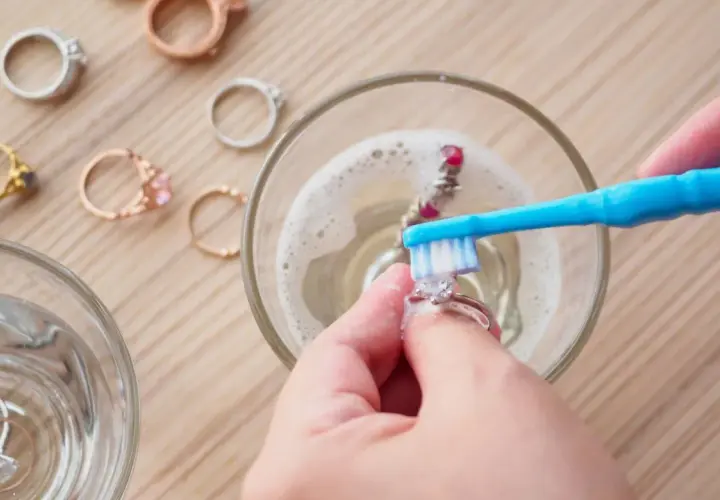 Manos limpian joyas con un cepillo azul en agua con espuma. Otros anillos esperan su turno.