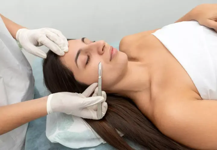Una esteticista realiza un dermaplaning facial a una mujer, eliminando vello fino y células muertas con un bisturí.