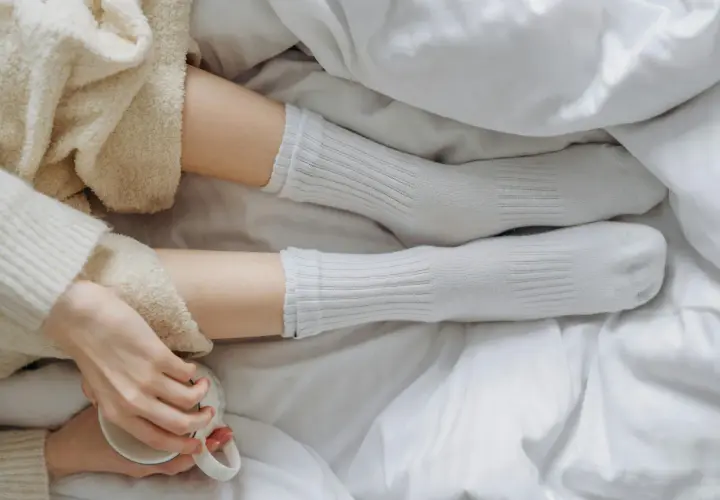 Piernas con calcetines blancos de poliamida, envueltas en mantas suaves, sosteniendo una taza.