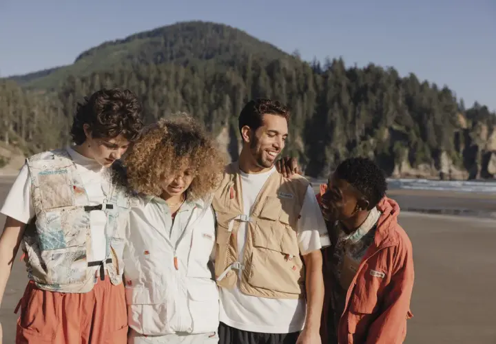 Grupo de amigos con estilo gorpcore en la playa, con chalecos utilitarios y chaquetas, disfrutando del paisaje costero.