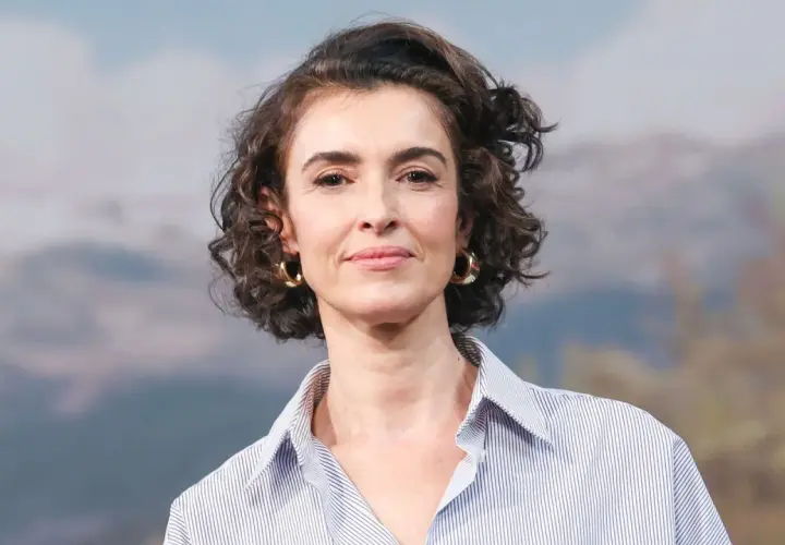 Blanca Romero, con su cabello rizado y pendientes dorados, sonríe con una camisa a rayas.