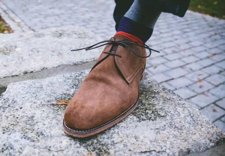 Zapato de gamuza marrón con cordones negros, calcetín a rayas y un consejo sobre como quitar manchas de humedad en zapatos de piel.
