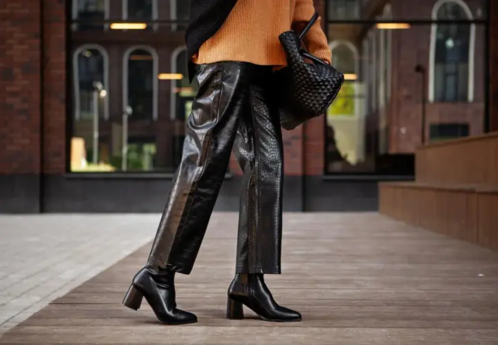 Elegante outfit con botas altas negras, pantalones de cuero texturizado y suéter naranja.