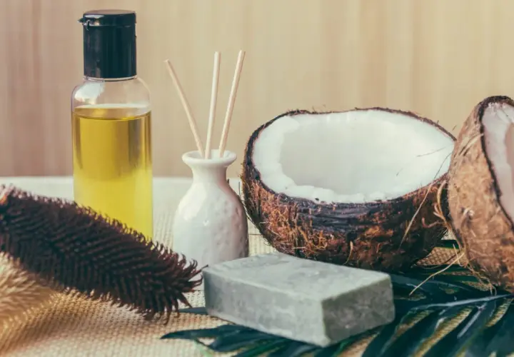 Aceite de coco para el pelo, junto a un coco partido, jabón y un cepillo.