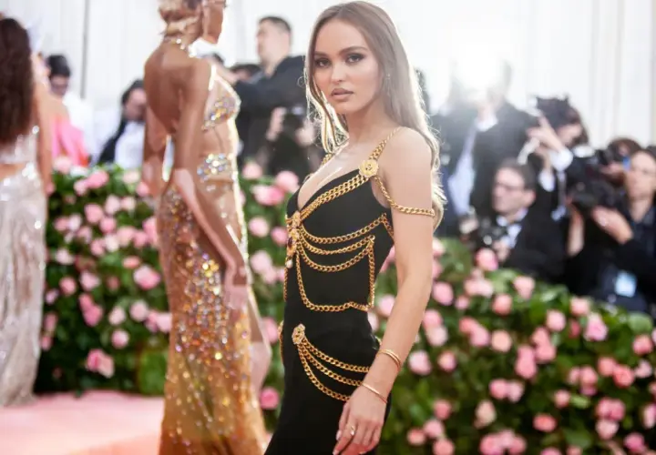 Lily-Rose Depp deslumbrante con un vestido negro adornado con cadenas doradas, posando ante un fondo de flores rosas y fotógrafos.
