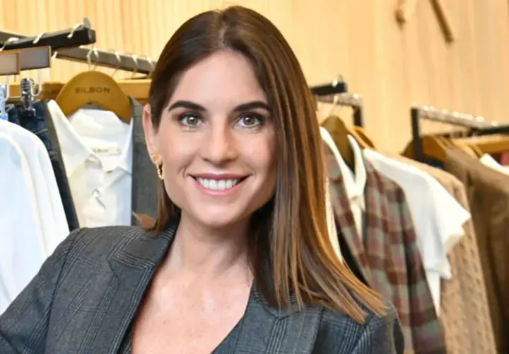 Lourdes Montes sonríe con un traje gris a cuadros, rodeada de ropa colgada en perchas en una tienda.