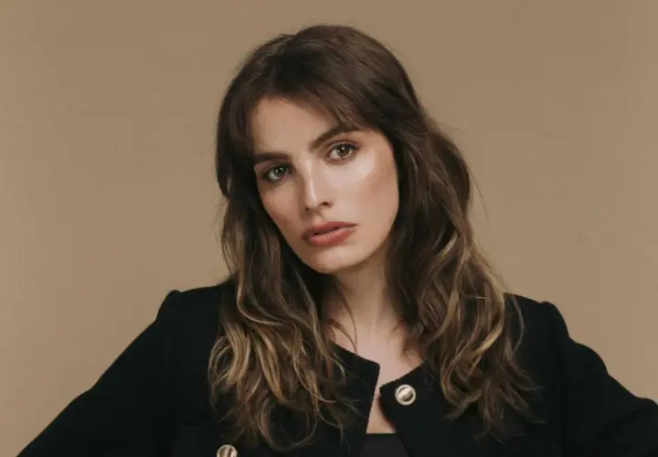 Retrato de una mujer con cabello castaño ondulado y flequillo, luciendo un blazer negro. Su mirada es intensa, transmitiendo un aire de misterio y elegancia, como si fuera la protagonista de una historia de lux pascal.