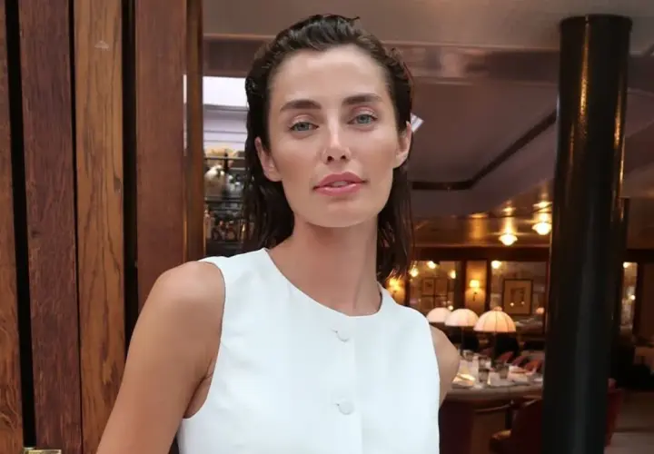 Rebecca Donaldson, con su mirada penetrante y labios rosados, posa con un elegante vestido blanco en un restaurante con ambiente cálido.