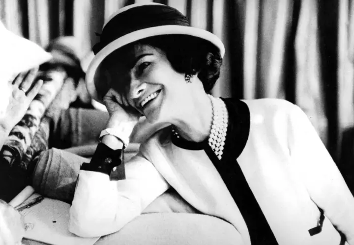 Coco Chanel, sonriendo con un sombrero y perlas, icónica figura de la moda.