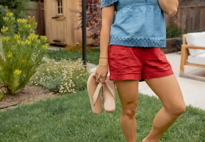 Mujer con blusa vaquera y shorts rojos, sosteniendo chanclas. El jardín tiene césped y flores.