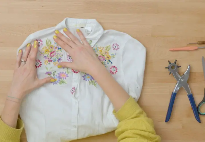 Manos embelleciendo una camisa blanca con bordados florales, lista para un proyecto de upcycling ropa. Herramientas de costura a un lado.