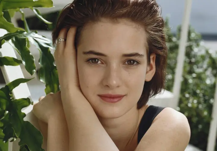 Winona Ryder joven, con su mano en la mejilla y un anillo discreto, rodeada de vegetación exuberante.