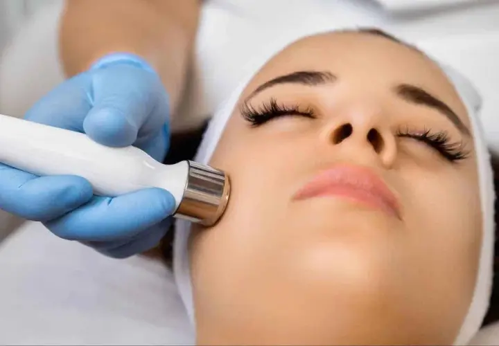 Un rostro recibe un tratamiento facial con exosomas, aplicado con un dispositivo de mano por un profesional con guantes azules.