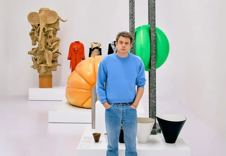 Jonathan Anderson posa entre esculturas y objetos de diseño, incluyendo una figura de madera tallada y un gran globo verde.