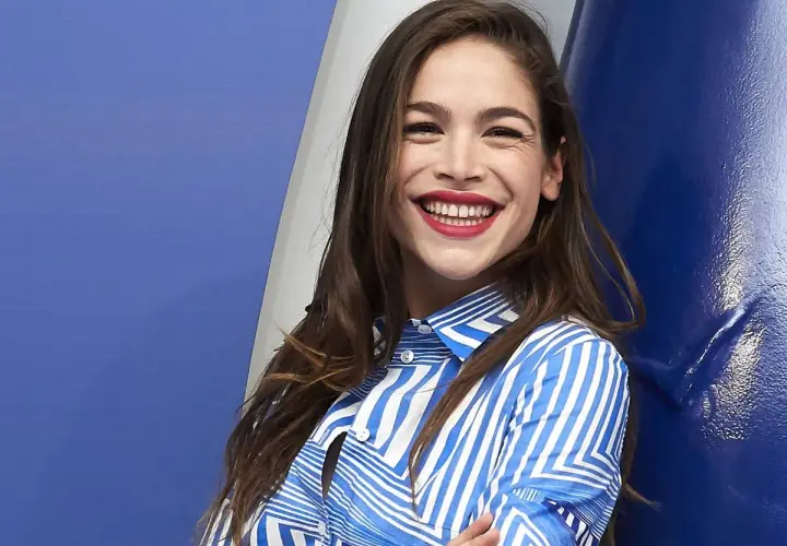 Cosima Ruiz de la Prada, sonriendo con labios rojos y camisa a rayas azules y blancas, se apoya en una pared azul.