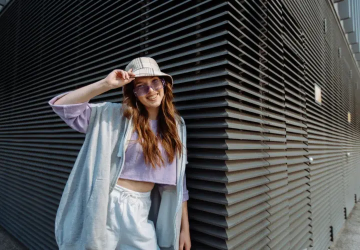 Chica vsco con sombrero y gafas de sol moradas, sonriendo y apoyada en una pared de lamas grises.