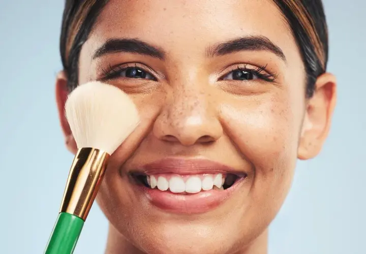 Una mujer sonríe mientras se aplica maquillaje con una brocha. Descubre trucos de maquillaje profesional para realzar tu belleza natural.