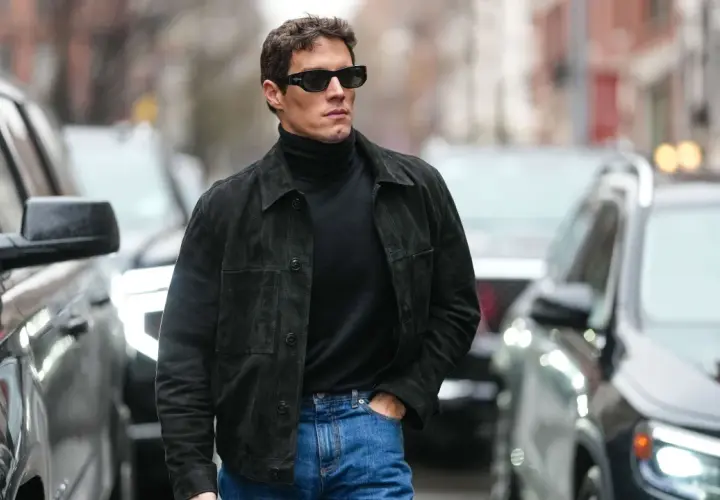 Hombre con estilo casual, viste jersey negro de cuello alto, chaqueta de ante y jeans. Camina por la calle entre coches.
